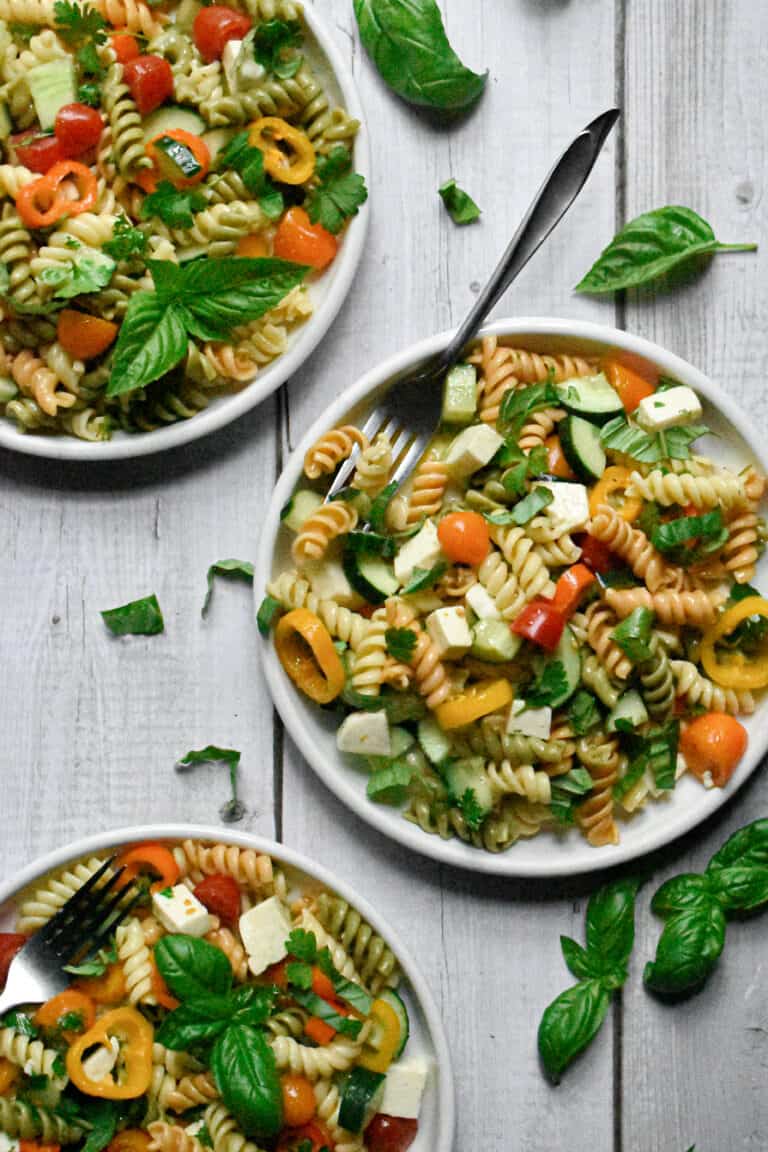 garden vegetable and herb pasta salad on three plates surrounded by fresh basil