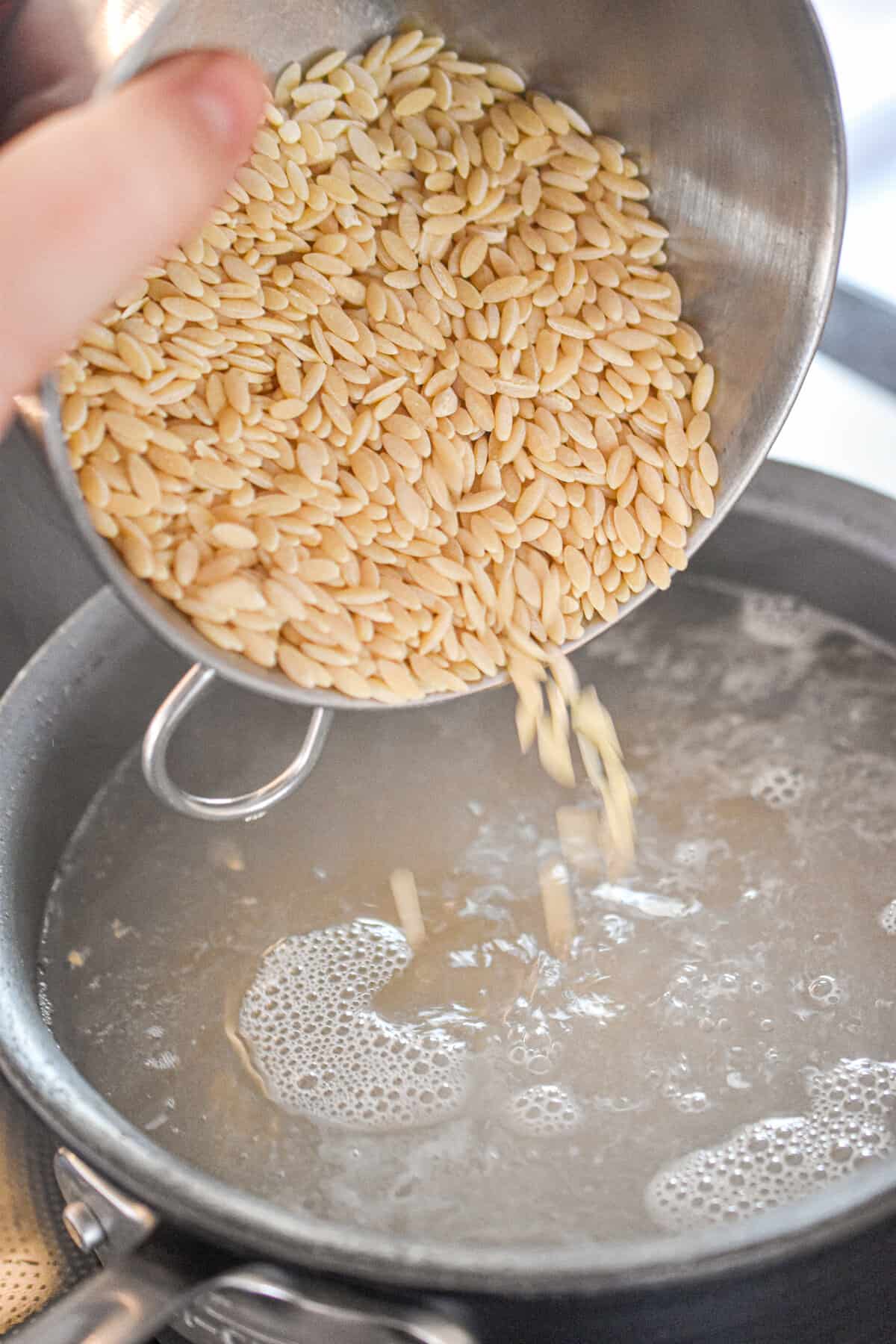 The orzo is being poured into the pot with the boiling water.
