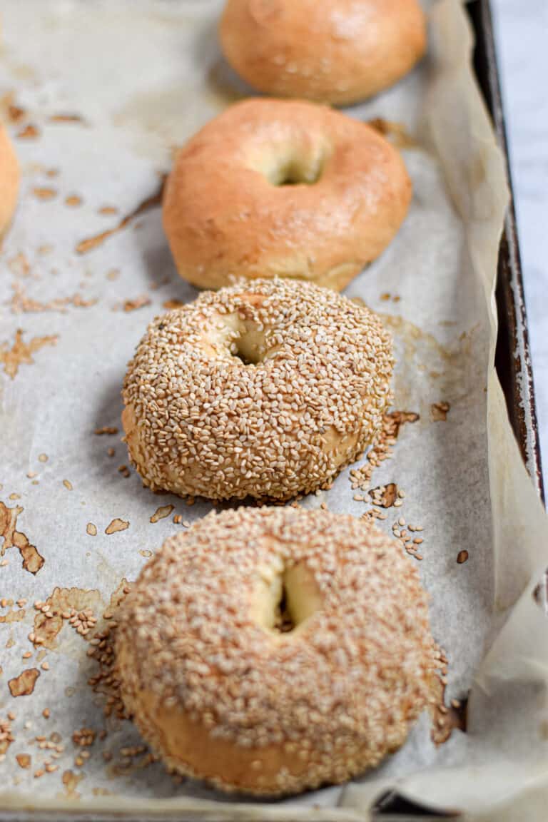 Two sesame seed Instant Pot bagels are on a baking sheet with two other Instant Pot bagels that are plain.