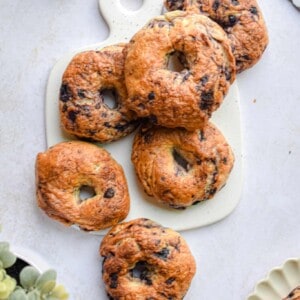 A bagel is on a plate and the rest of the blueberry bagels are on a cutting board.