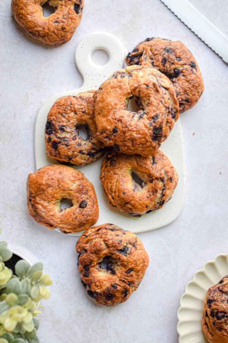 A bagel is on a plate and the rest of the blueberry bagels are on a cutting board.