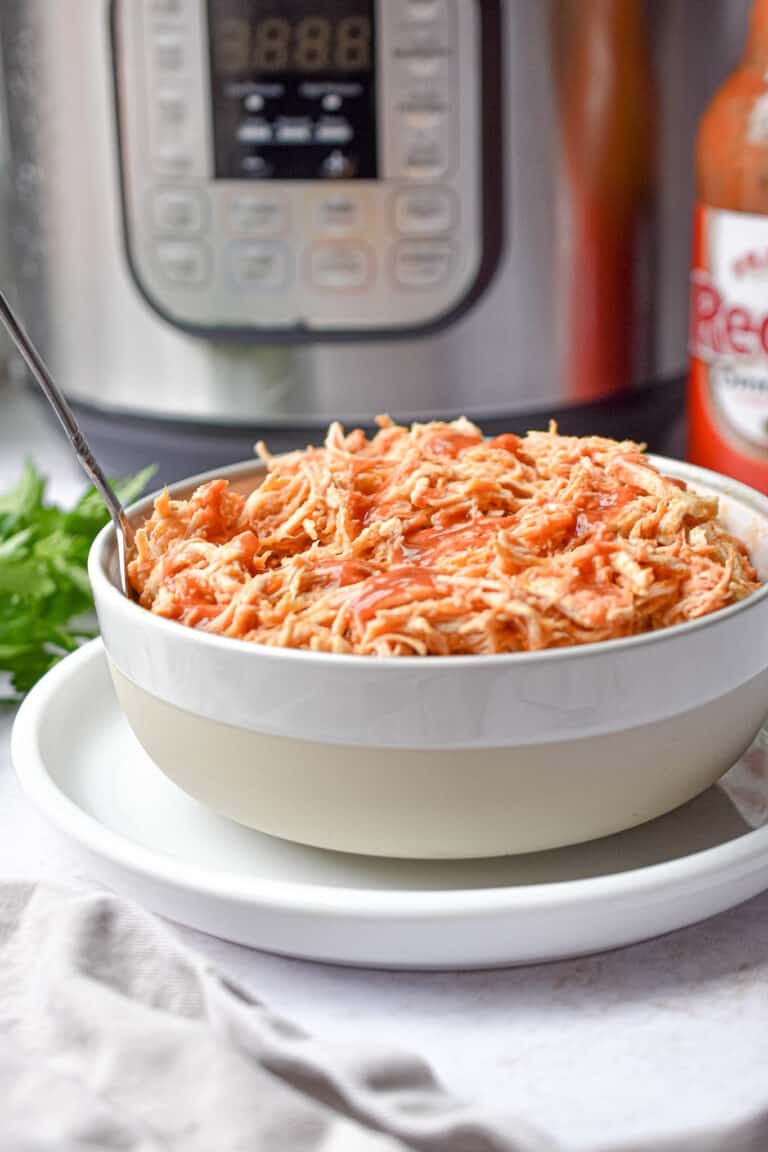 A bowl of buffalo chicken is in front of an Instant Pot and hot sauce.