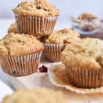 A pile of cranberry walnut muffins is in front of an unwrapped muffin.