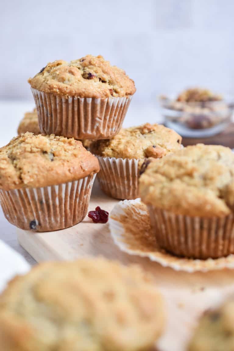 A pile of cranberry walnut muffins is in front of an unwrapped muffin.