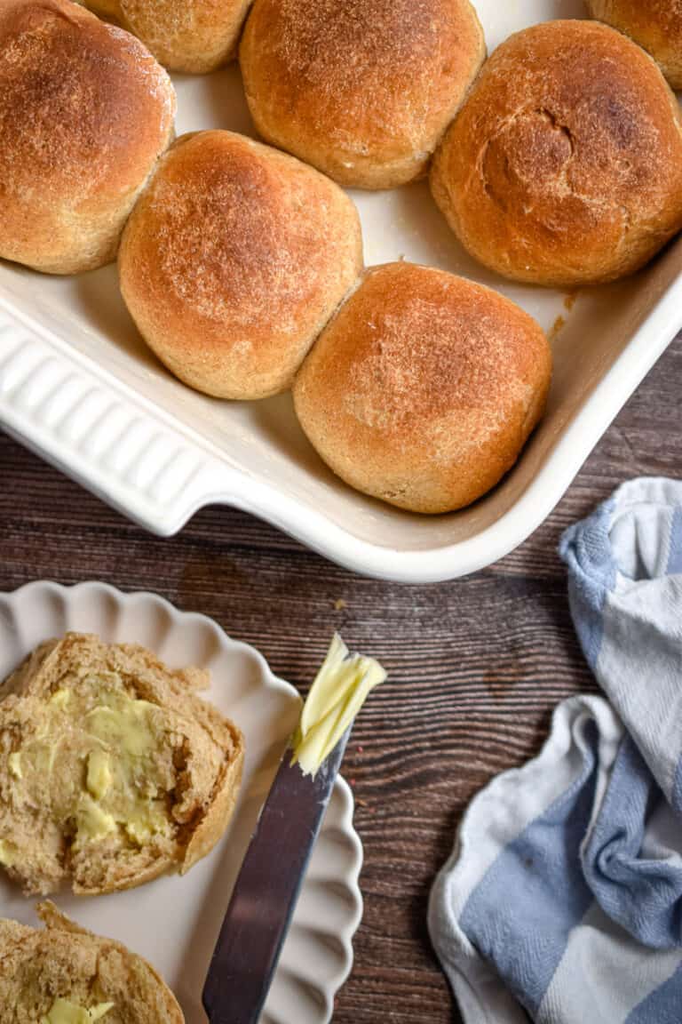 A whole wheat roll is cut open with butter on a knife and the rest of the rolls in a white baking dish.