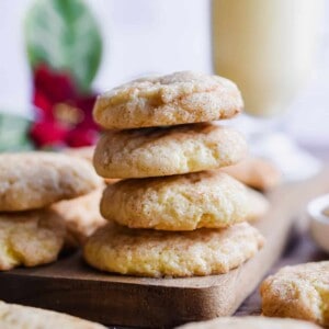 Four cookies are stacked on a cutting board with eggnog in the background.