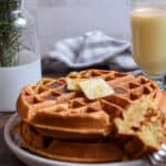 The eggnog waffles are on a plate with a glass of eggnog in the background with pine twigs in a vase.