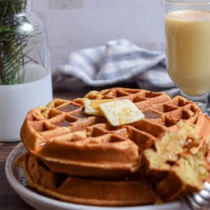 The eggnog waffles are on a plate with a glass of eggnog in the background with pine twigs in a vase.