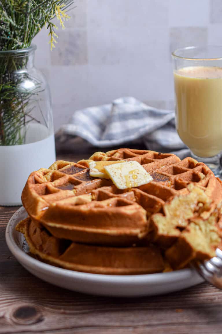The eggnog waffles are on a plate with a glass of eggnog in the background with pine twigs in a vase.