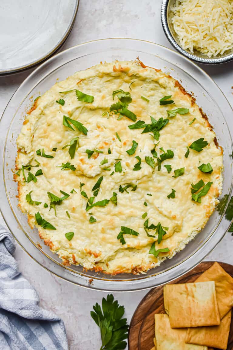 The dip is in a clear baking dish surrounded by pita chips and parsley.