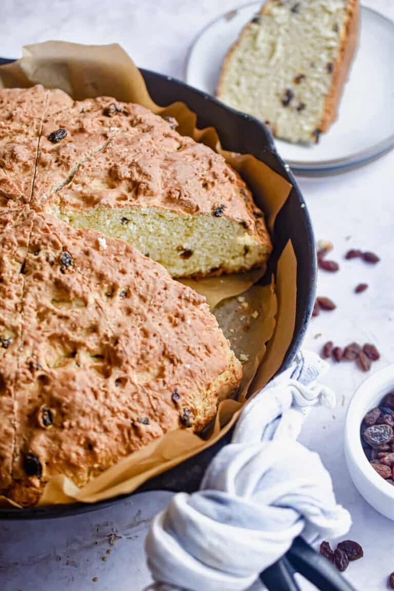 A piece of sourdough irish soda bread is on a plate next to the cast iron skillet.
