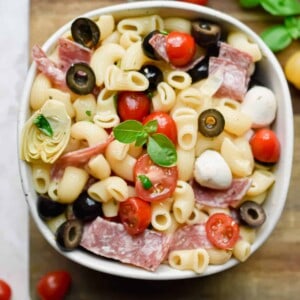 A bowl of the Italian Antipasto Pasta Salad is on a cutting board and surrounded by tomatoes and basil.