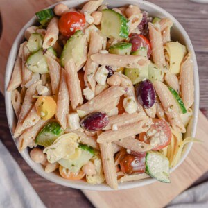 A bowl of Greek Pasta Salad with Penne Pasta is sitting on a cutting board.