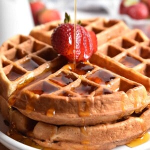 Maple syrup is being poured on a stack of strawberry waffles.