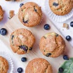 A group of banana blueberry muffins are on a cutting board with leaves and blueberries around them.