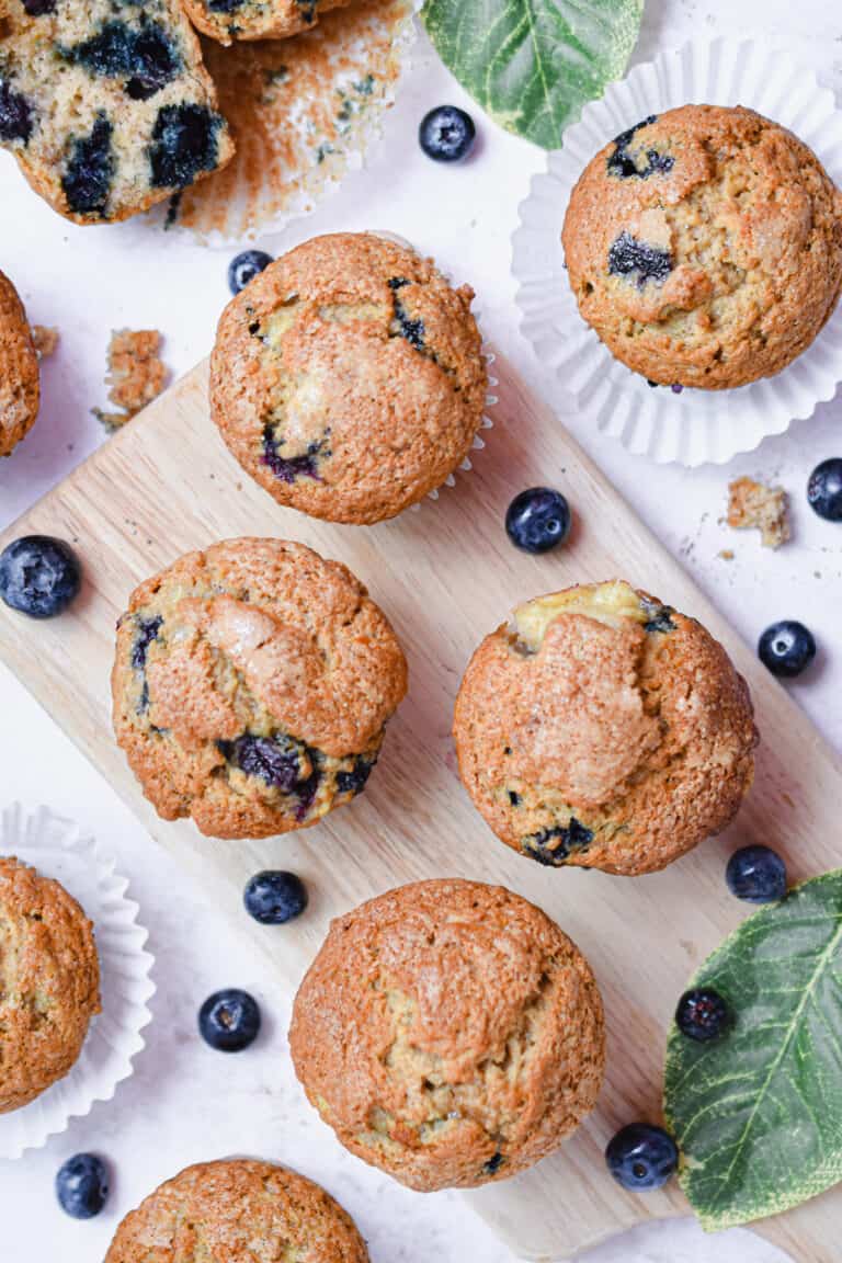 A group of banana blueberry muffins are on a cutting board with leaves and blueberries around them.
