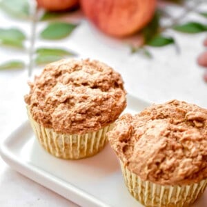 Two 3 ingredient apple spice muffins are on a square plate with apples in the back.