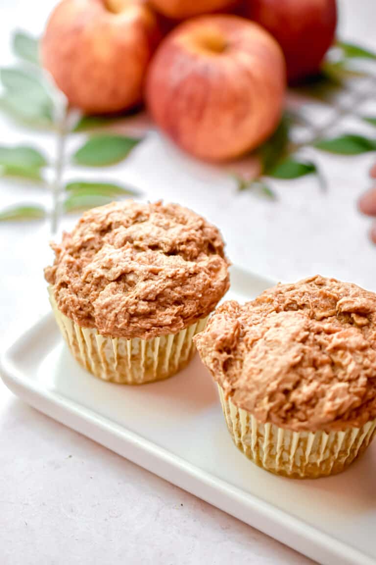 Two 3 ingredient apple spice muffins are on a square plate with apples in the back.