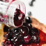 A jar of the blueberry sauce for pancakes is being poured over the pancakes.