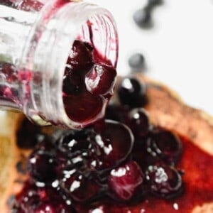 A jar of the blueberry sauce for pancakes is being poured over the pancakes.