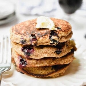 A stack of blueberry oatmeal pancakes have a glass of coffee behind them.