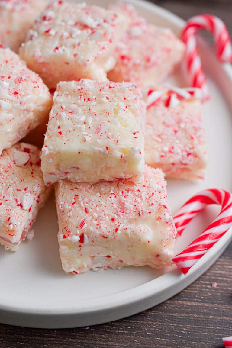 A pile of white chocolate peppermint fudge is on a white plate surrounded by small candy canes.
