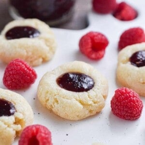 One 5 ingredient raspberry cheesecake thumbprint cookies are surrounded by raspberries and jam.