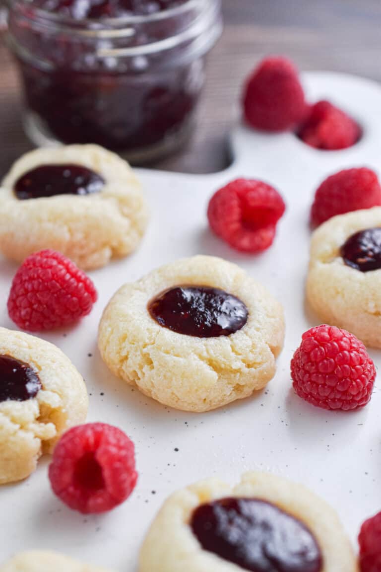 One 5 ingredient raspberry cheesecake thumbprint cookies are surrounded by raspberries and jam.