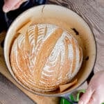 Two hands are holding a blue Dutch oven with a loaf of sourdough bread in it.