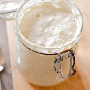 The sourdough starter is bubbly and in a glass jar on a cutting board.