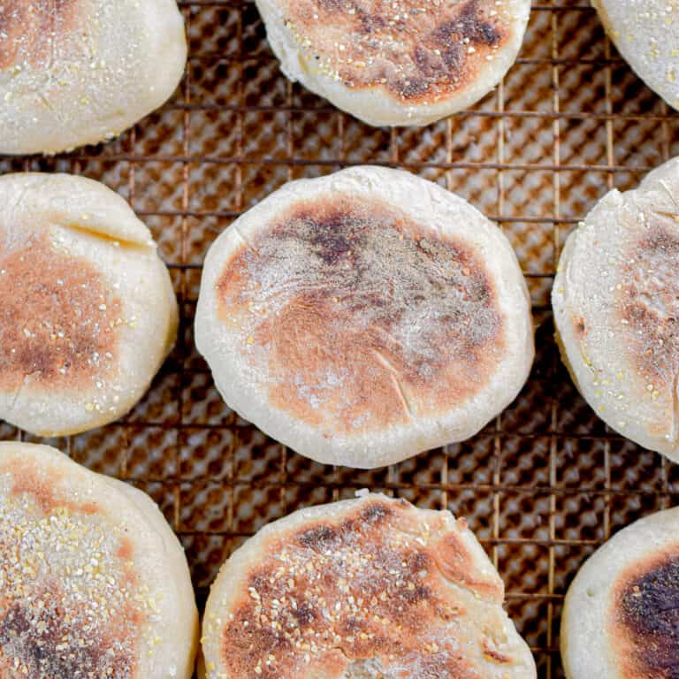 Nine sourdough English muffins are laid out on a wire rack over a baking sheet.
