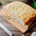 The sourdough discard focaccia is on a wooden cutting board with a serrated knife in front of it.