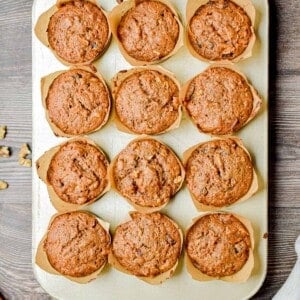 12 sourdough morning glory muffins are in a muffin tin surrounded by nuts, carrots, and apples.