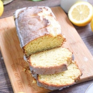 A sourdough discard lemon loaf is on a wooden cutting board with a plant in the back and surrounded by cut lemons.