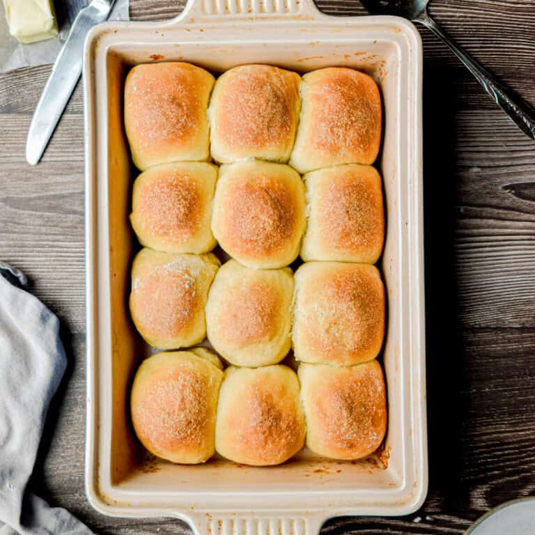 Twelve sourdough Hawaiian rolls are in a baking dish with a butter knife and a towel around the baking dish.