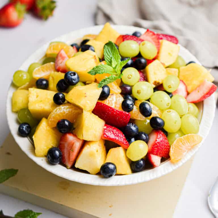 An overflowing bowl of fruit salad has a sprig of mint on it with fresh strawberries in the background.