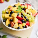 A bowl of fruit salad is on a square platter with min sprigs around the bowl.