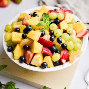 A bowl of fruit salad is on a square platter with min sprigs around the bowl.