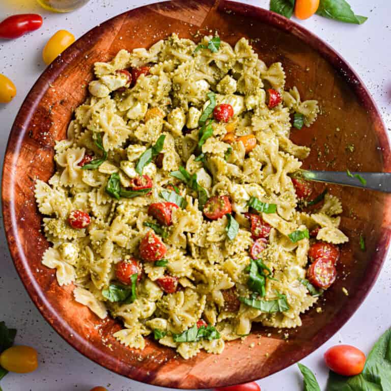 The pesto pasta salad is in a large wooden bowl surrounded by basil and tomatoes.