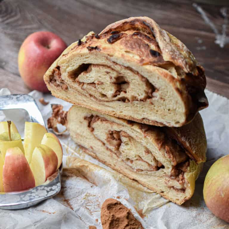 Apples are in the background of two halves of the apple cinnamon sourdough bread stacked on top of each other.