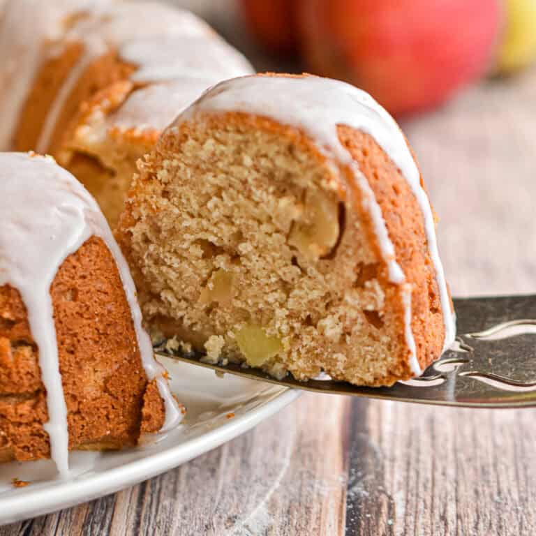 The sourdough apple cake is topped with powdered sugar icing and a slice is being taken out of the cake.