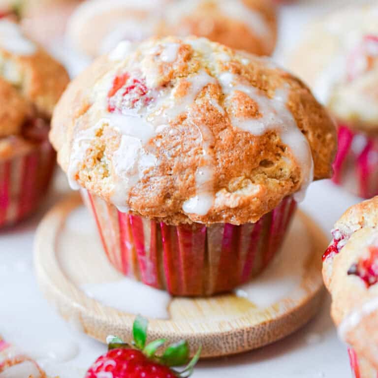 Icing is on top of the sourdough strawberry muffin surrounded by muffins and strawberries.
