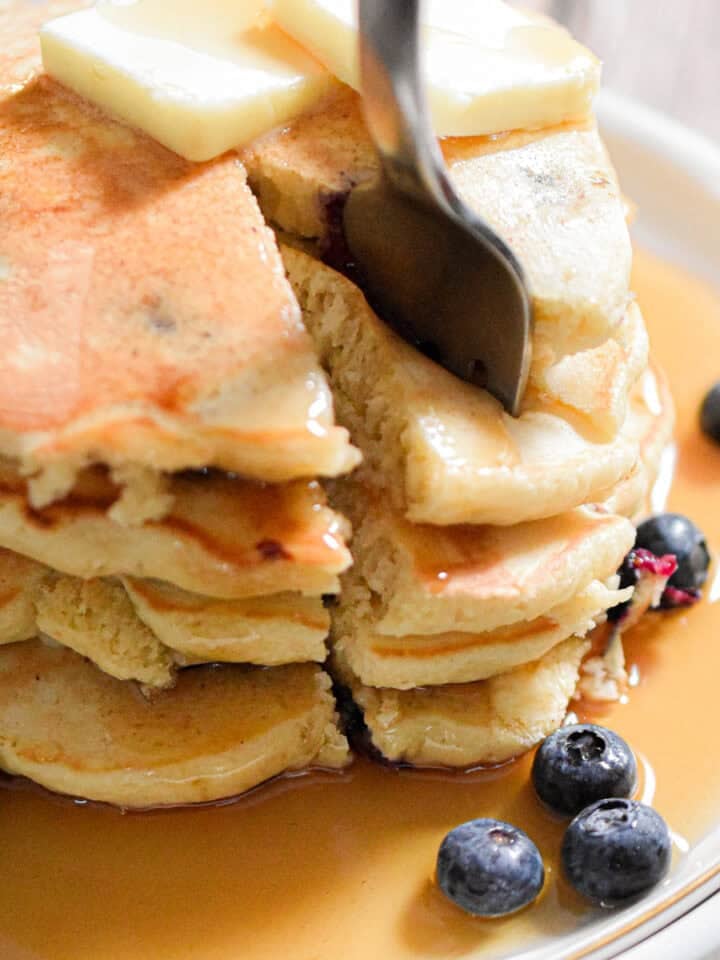 A fork is in a portion of the blueberry Bisquick pancakes on a plate with maple syrup and blueberries on it.