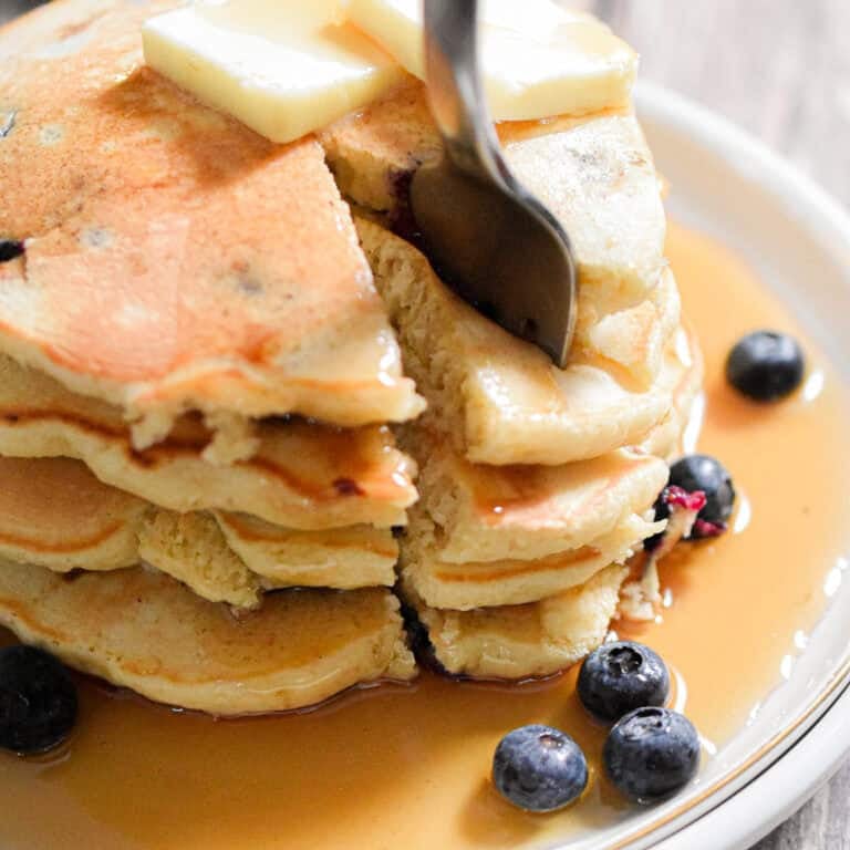 A fork is in a portion of the blueberry Bisquick pancakes on a plate with maple syrup and blueberries on it.