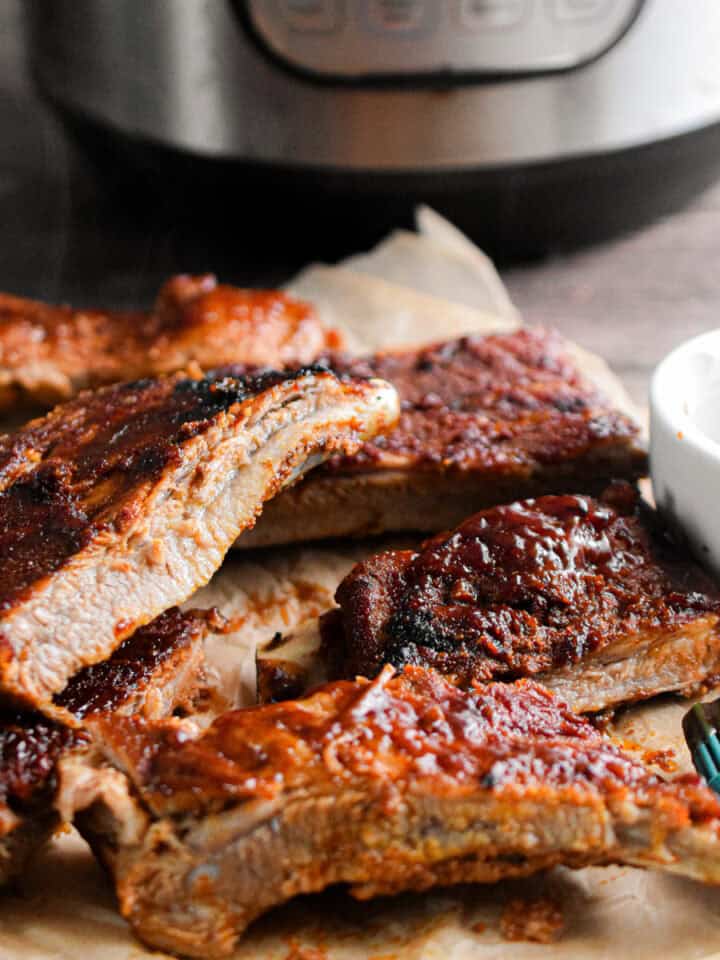 The ribs are piled in front of the Instant Pot with a bowl of barbecue sauce next to them.