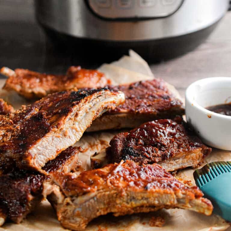 The ribs are piled in front of the Instant Pot with a bowl of barbecue sauce next to them.