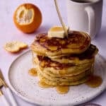 A half of a peeled orange is behind a plate with the cornmeal pancakes being drizzled with maple syrup.
