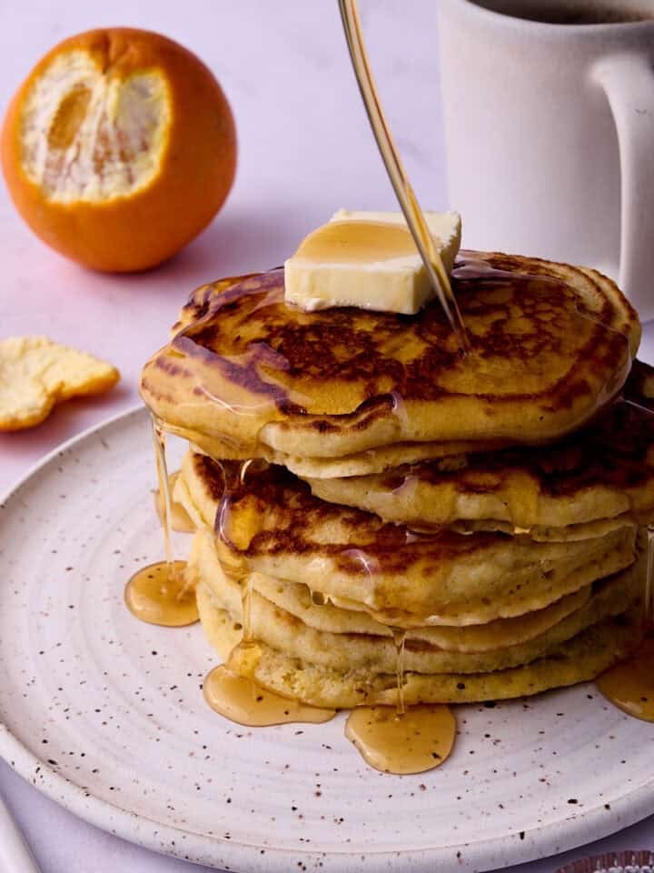 A half of a peeled orange is behind a plate with the cornmeal pancakes being drizzled with maple syrup.