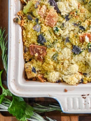 Fresh rosemary and fresh sage are next to a baking dish on a wooden cutting board.