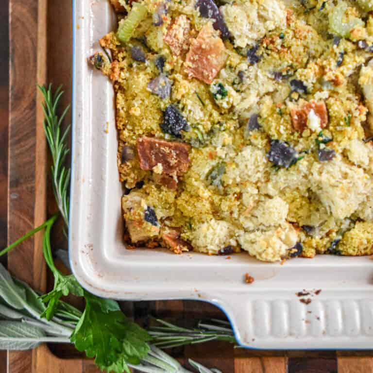 Fresh rosemary and fresh sage are next to a baking dish on a wooden cutting board.